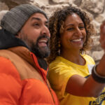 Two people with South Asian heritage take a selfie. One is a man with a beard wearing a bobble hat and orange puffer jacket, the other is a woman with a yellow t-shirt and curly hair.