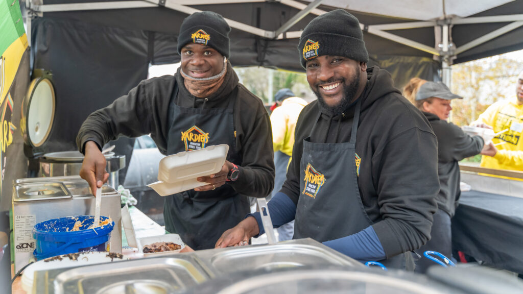 Two Black chefs smile at the camera as they serve the food they are cooking.