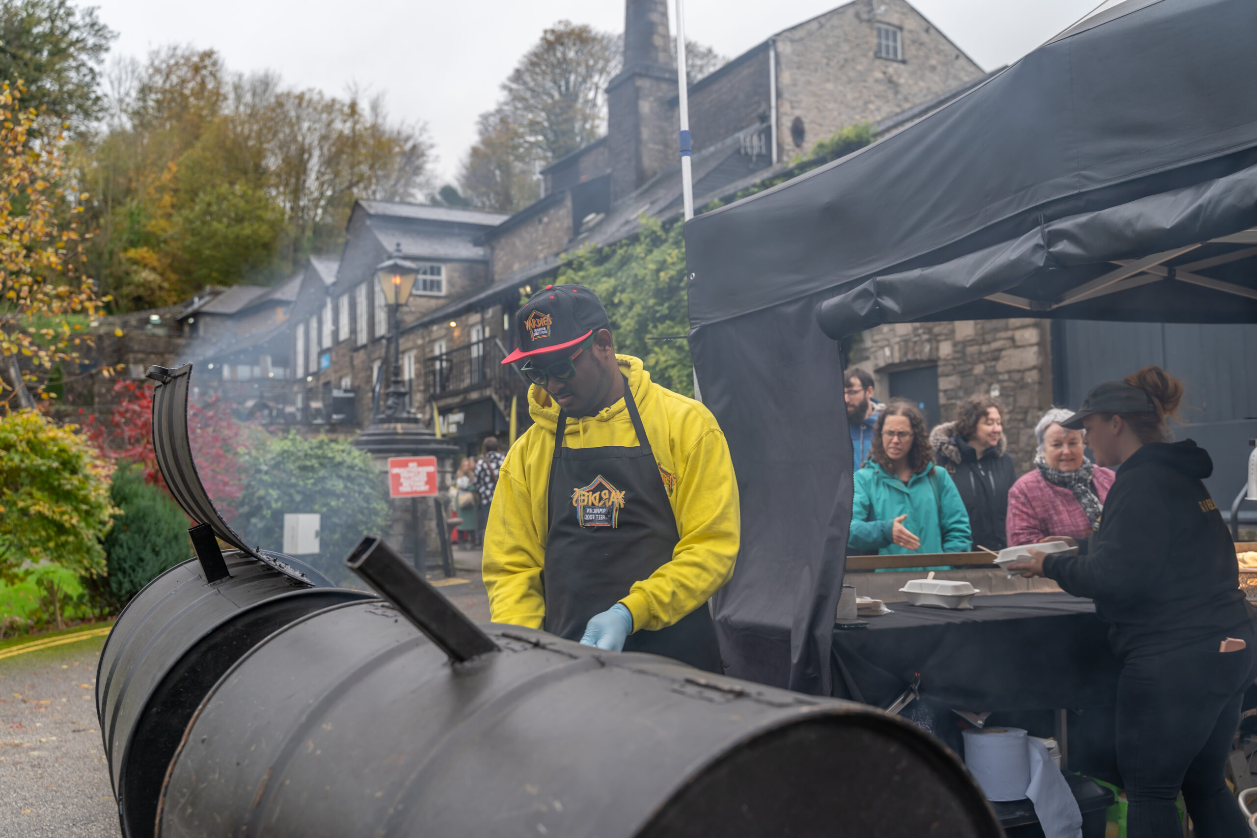 A Black man wearing a yellow top cooks in a tin barrel barbecue, smoke rises. In the background people are queuing to collect their food. 