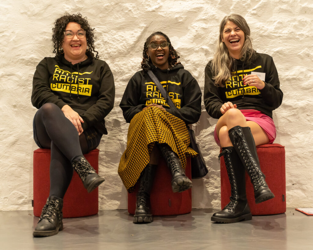 Three seated women, laugh and smile at the camera. Two of them are white the woman in the middle is Black.