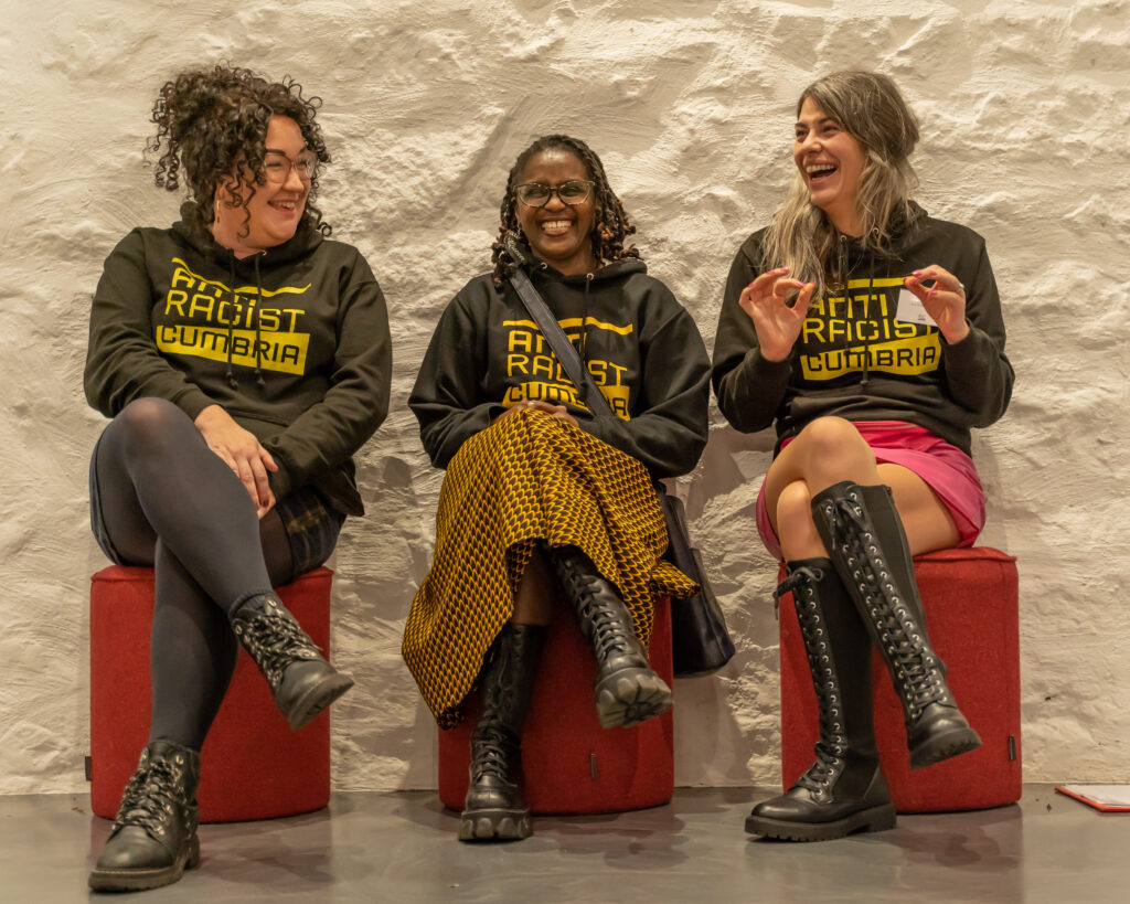 Three seated women, laugh and smile at each other. Two of them are white the woman in the middle is Black.