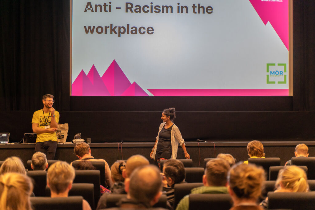 A woman with South Asian heritage and a white man talk at the front of a theatre of people.