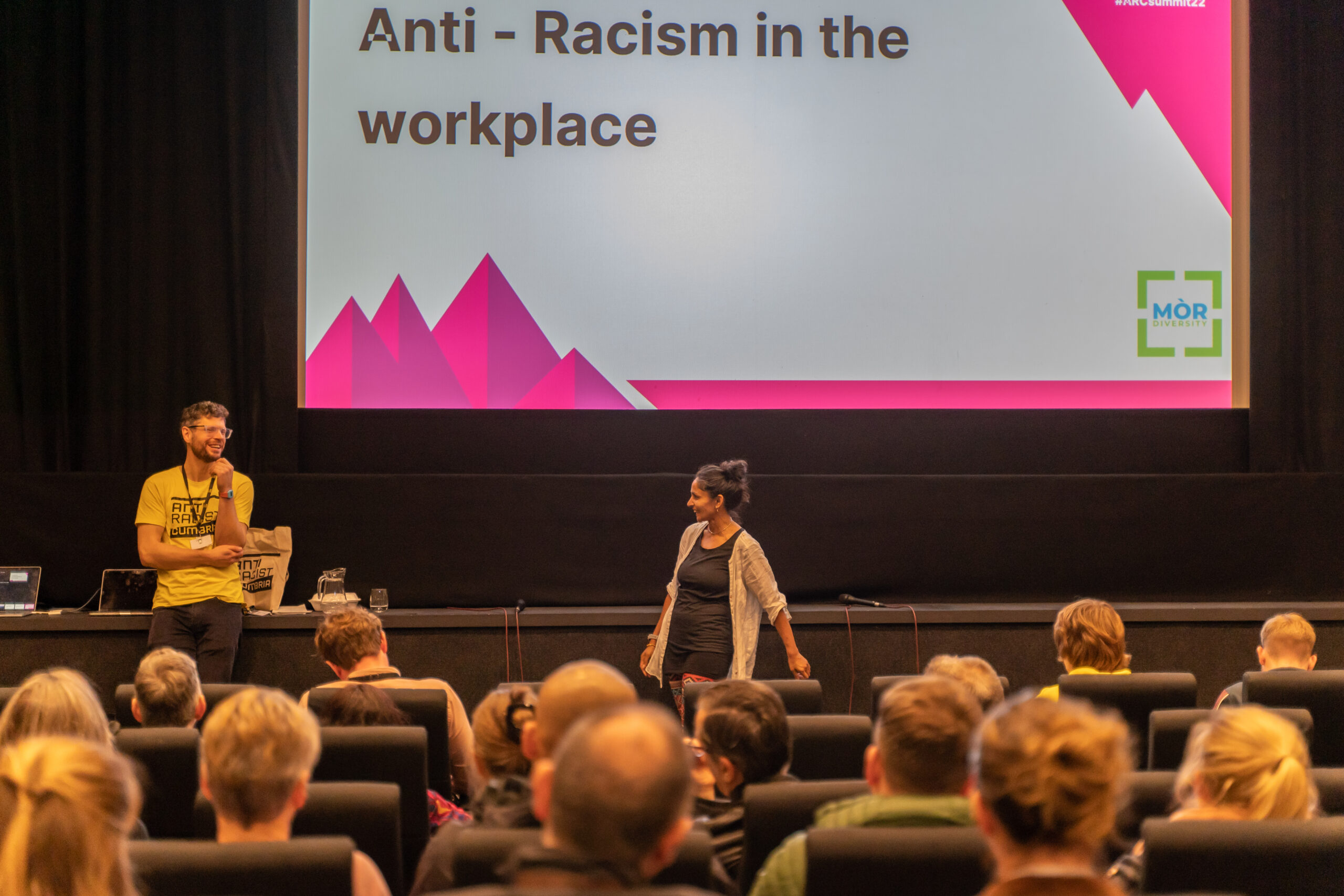 A woman with South Asian heritage and a white man talk at the front of a theatre of people.