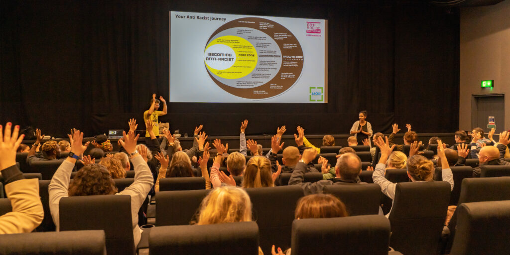 A woman with South Asian heritage and a white man talk at the front of a theatre of people. The audience wave their hands in appreciation.