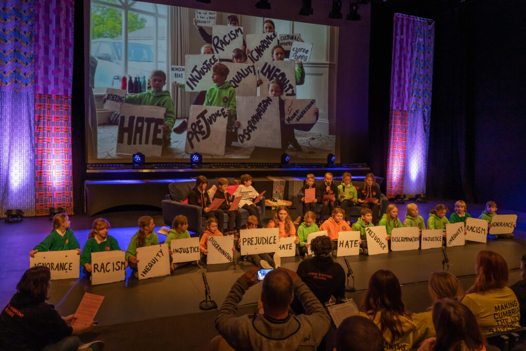 A row of primary school aged children sit on stage. They each hold a placard with words such as 'injustice' 'inequity' 'hate' and 'prejudice'