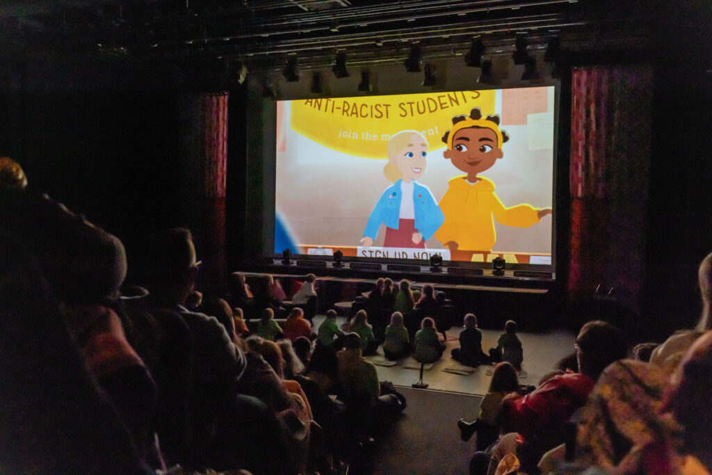 A screen with a shot from animated film The Fell We Climb is on the screen, in front a full theatre watches.