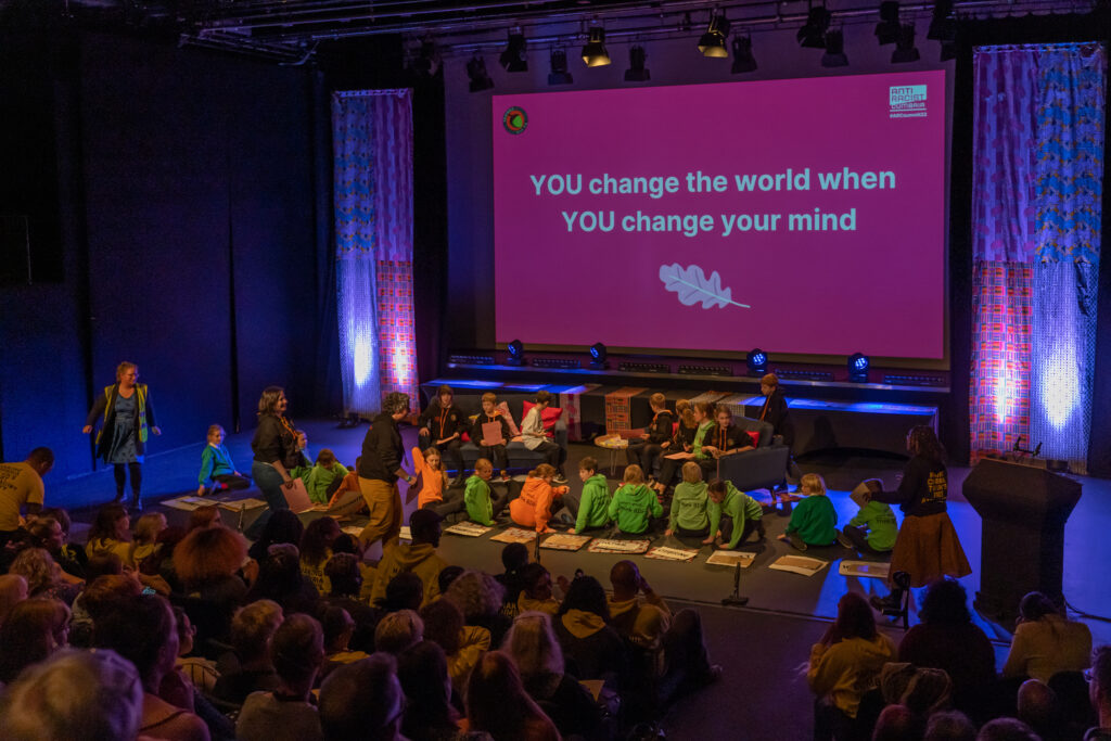 A screen with wording saying 'you change the world when you change your mind' is shown in front a full theatre watches.