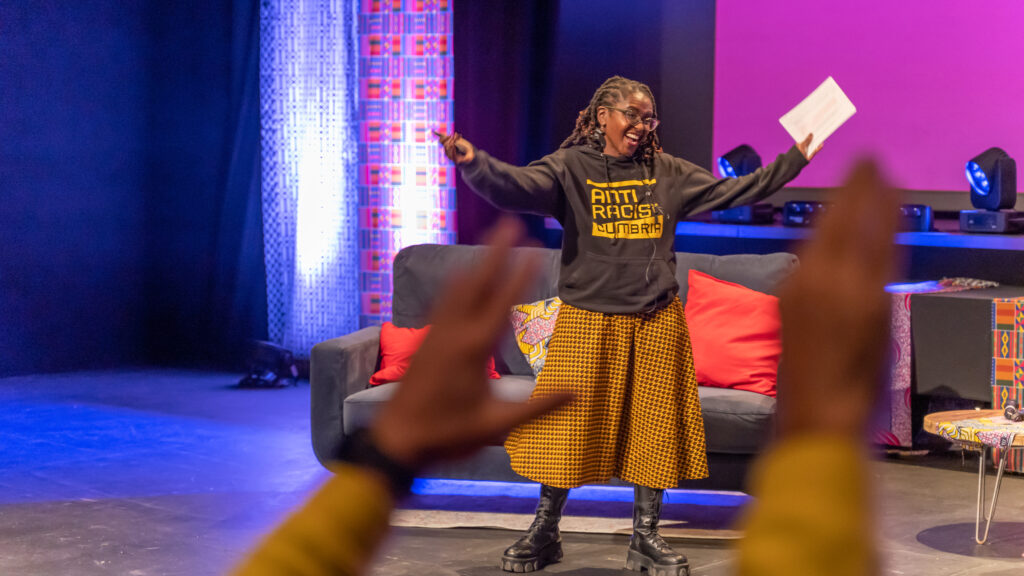 A Black woman talks on stage her arms waving. The audience are also waving their hands. She wears an Anti Racist Cumbria hoody and an African print skirt. Her locs are twisted up to a shoulder length.