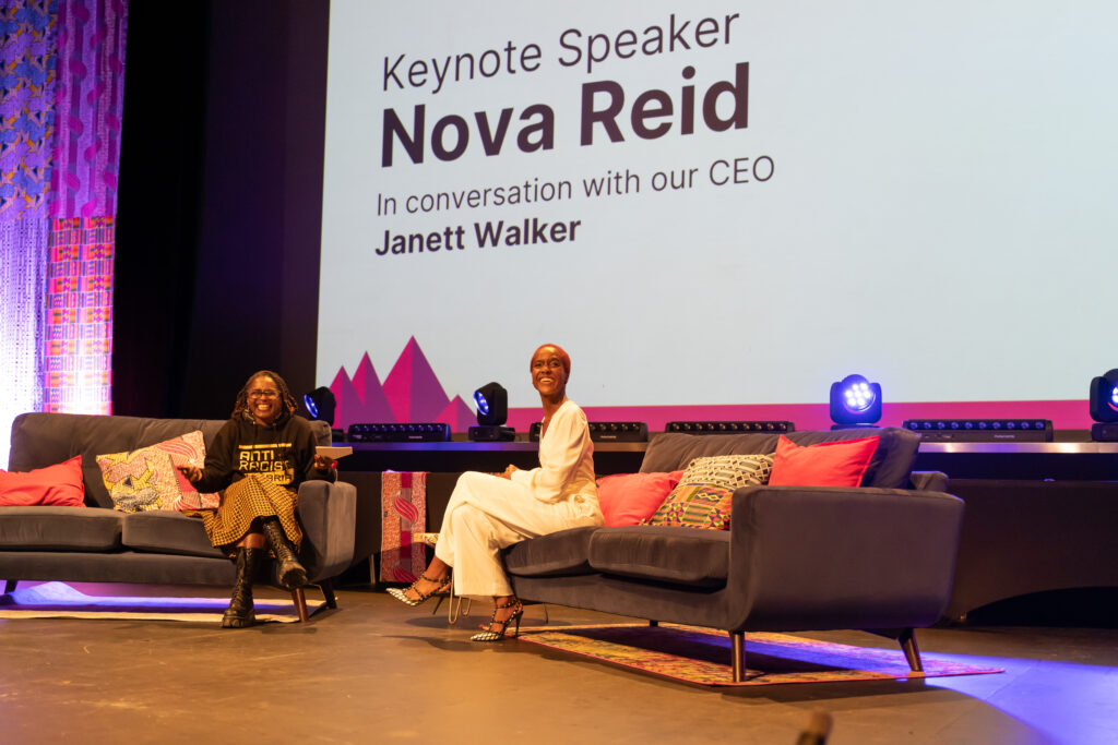 Two Black sit on a different sofa each, they are both smiling. The audience are also waving their hands. The woman on the left wears an Anti Racist Cumbria hoody and an African print skirt. Her locs are twisted up to a shoulder length. The woman on the right has short red afro hair, she wears white trousers and white top.