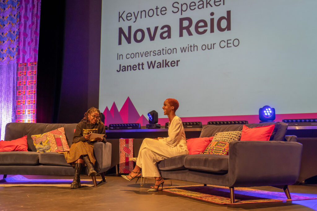Two Black sit on a different sofa each, they are both smiling. The audience are also waving their hands. The woman on the left wears an Anti Racist Cumbria hoody and an African print skirt. Her locs are twisted up to a shoulder length. The woman on the right has short red afro hair, she wears white trousers and white top.