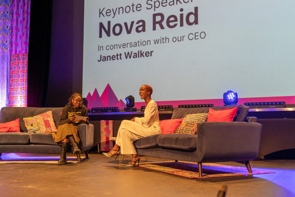 Two Black sit on a different sofa each, they are both smiling. The audience are also waving their hands. The woman on the left wears an Anti Racist Cumbria hoody and an African print skirt. Her locs are twisted up to a shoulder length. The woman on the right has short red afro hair, she wears white trousers and white top.