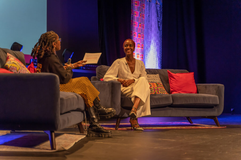 Two Black sit on a different sofa each, they are both smiling. The audience are also waving their hands. The woman on the left wears an Anti Racist Cumbria hoody and an African print skirt. Her locs are twisted up to a shoulder length. The woman on the right has short red afro hair, she wears white trousers and white top.