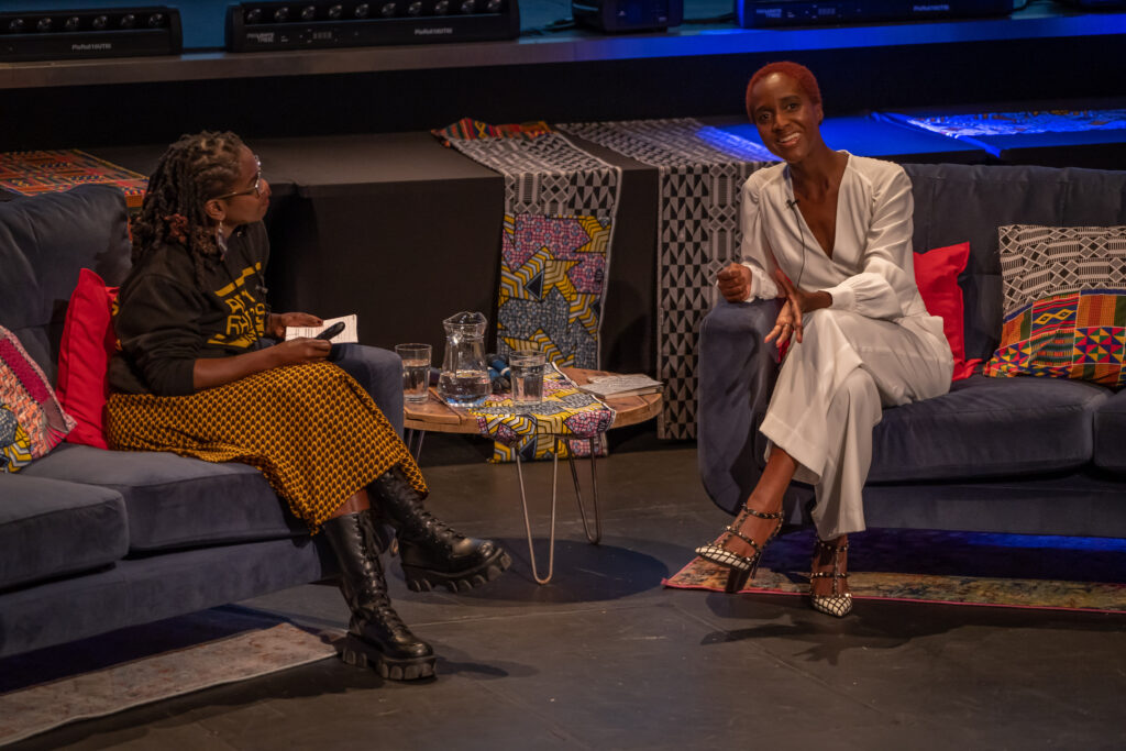 Two Black women sit on a different sofa each, they are both smiling. The audience are also waving their hands. The woman on the left wears an Anti Racist Cumbria hoody and an African print skirt. Her locs are twisted up to a shoulder length. The woman on the right has short red afro hair, she wears white trousers and white top.