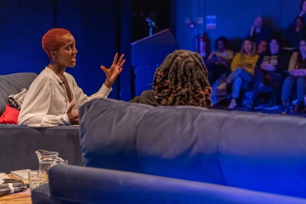 A Black woman wearing white is talking animatedly, over another Black woman's shoulder the audience looks on.