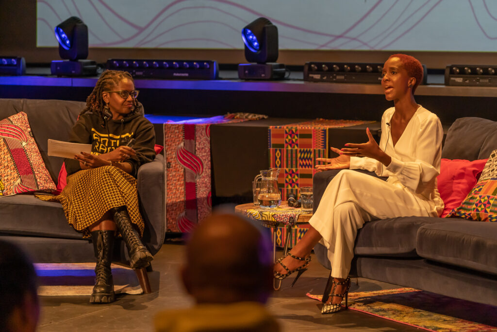 Two Black women converse as they sit on stage.