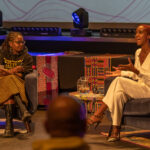 Two Black women converse as they sit on stage.