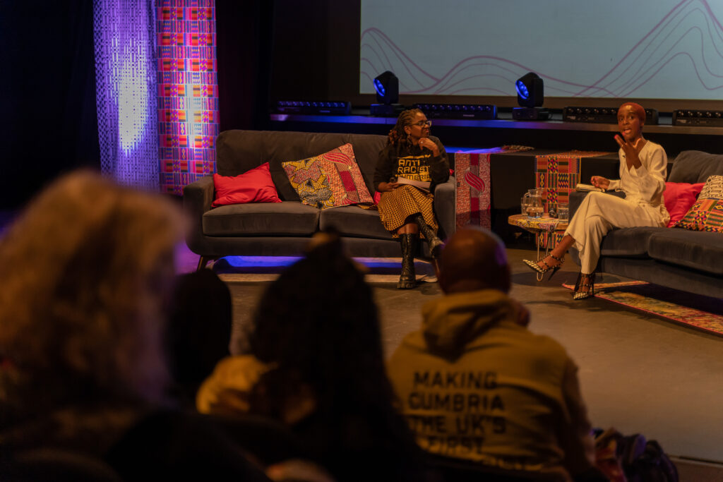 Two Black women converse as they sit on stage.