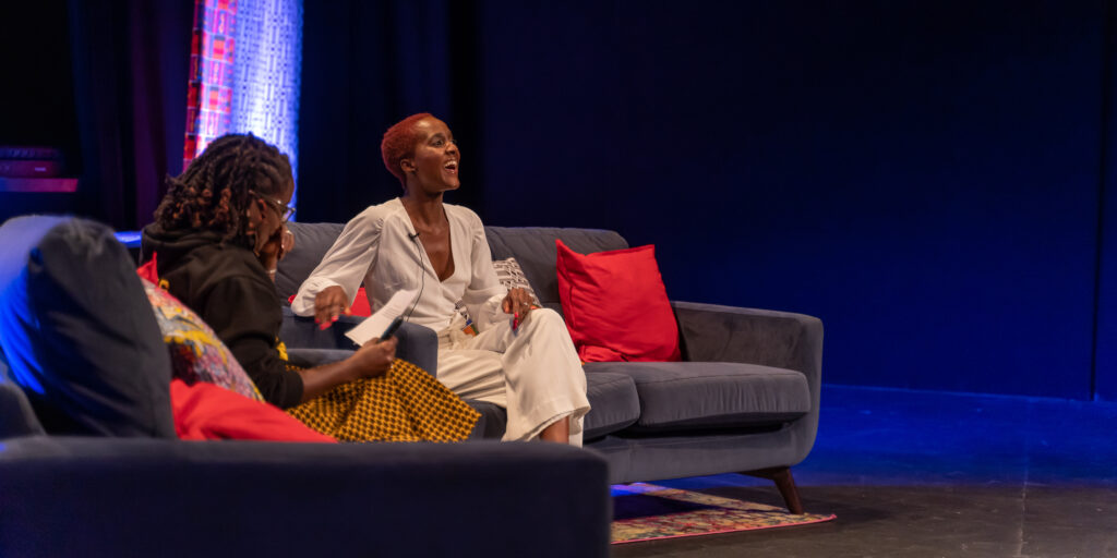 Two Black women converse as they sit on stage.