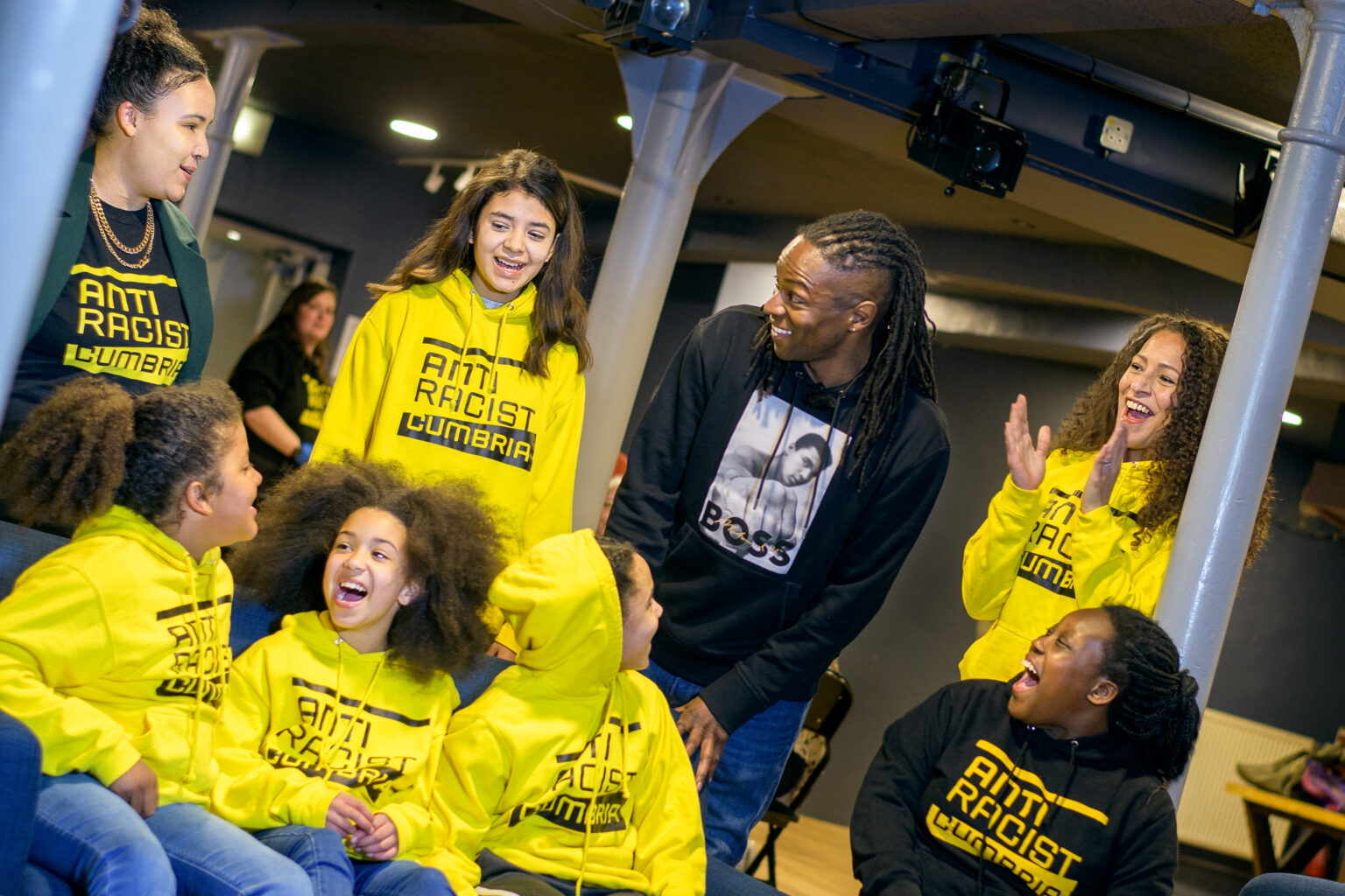 A group of young Black and Brown people from our youth grass roots project, all wearing yellow Anti Racist Cumbria Hoodies and laughing