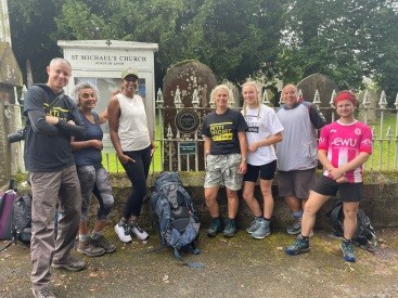 A group of smiling people stand by a plaque in a graveyard. The plaque commemorates the first recorded African community in Britain.