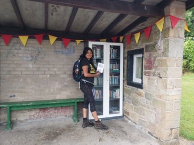 A woman stands inside a stone built bus shelter with a roof. There is red and yellow bunting around the ceiling, a green bench and a white painted bookshelf. The woman holds a book called Why I No Longer Talk to White People About Race.