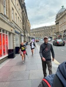People walking along a pavement. Tall buildings on the left loom over the walkers.