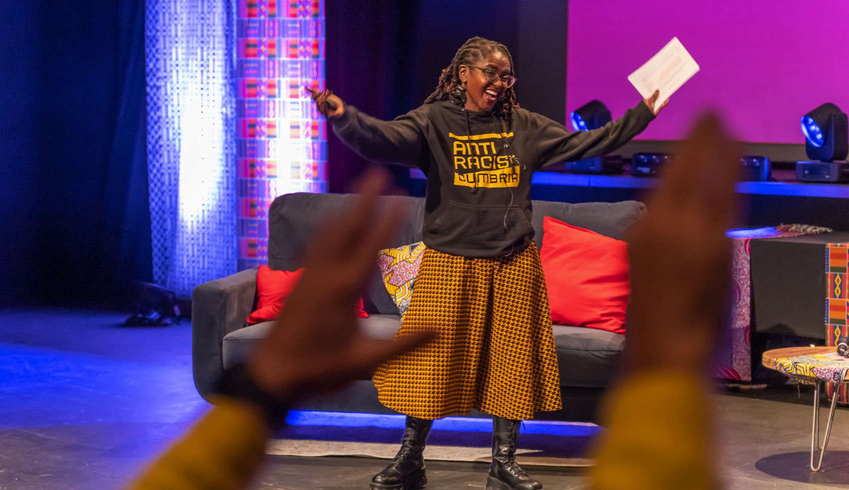Janett, the CEO of Anti-Racist Cumbria stands on stage at the ARC Summit with arms out wide, wearing an ARC hoodie and long orange skirt. Janett is a short black woman.