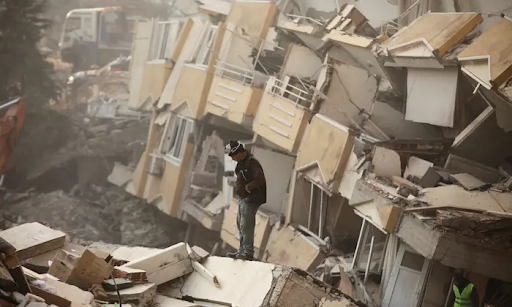 A man stands in front of destroyed buildings that are leaning precariously.