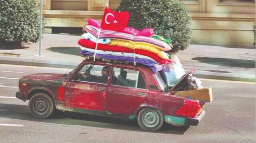A red car flying a Turkish flag, there are multiple mattresses and blankets strapped to the roof and the boot is overflowing with boxes