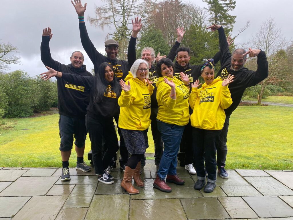 A group of 9 people of different genders, ages and ethnicities, all wearing Anti Racist Cumbria hoodies smiling and waving.