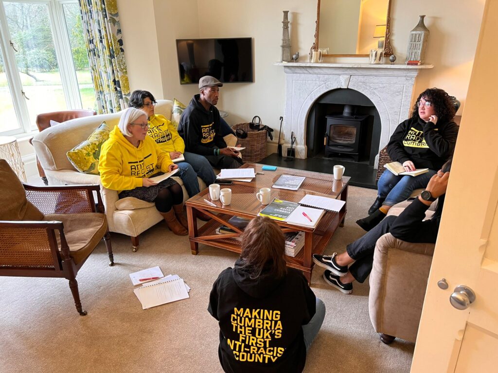 6 people wearing Anti Racist Cumbria hoodies sit on sofas in a sunlight room. They are talking animatedly.