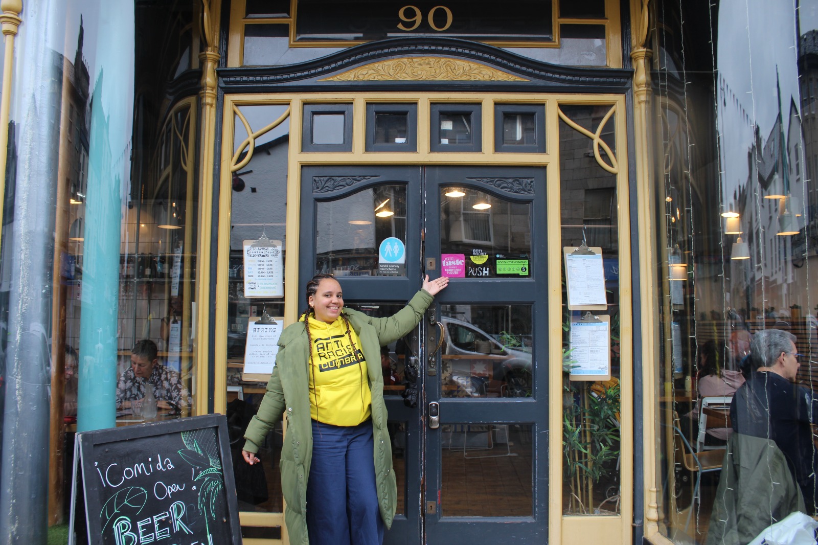 A young black woman stands in front of a bar wearing an Anti Racist Cumbria hoodie, showcasing an ARC sticker in the window.