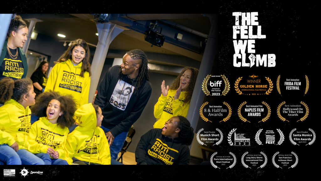 Promotional banner for The Fell We Climb film. Group of black and brown children wearing Anti Racist Cumbria hoodies with Nigel Clarke, a black man. All are smiling.