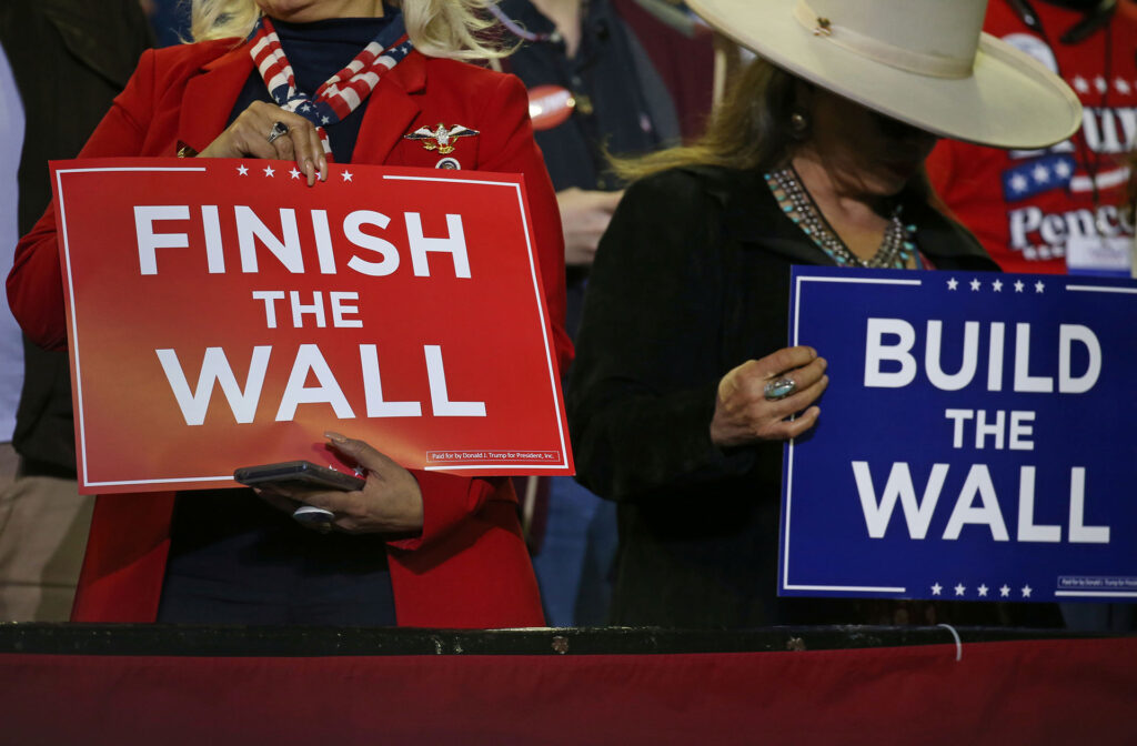 Two people hold political signs up. 1: Finish the Wall. 2. Build the Wall. 