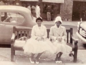 Two Black women wearing white dresses sit on a bench. It is the 1950s and the photo is in black and white.