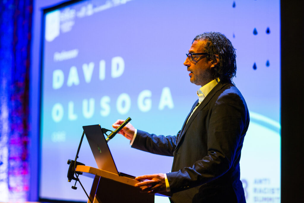David Olusoga addresses an audience. He is a Black man with shoulder length locs, he wears glasses and dark suit.