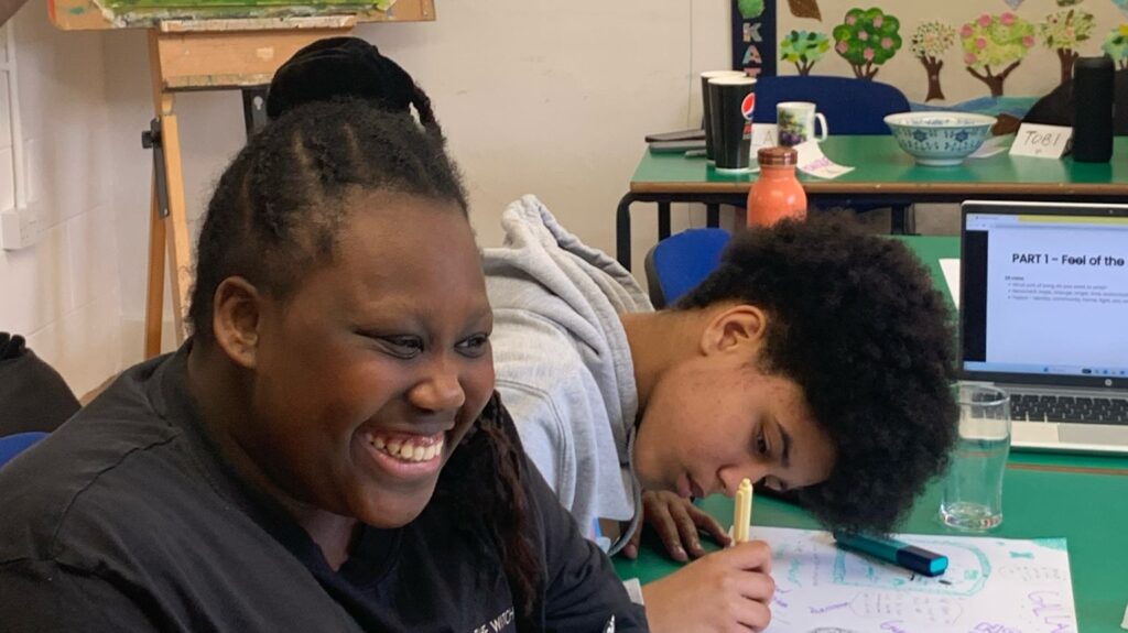 Two young black people drawing at a table. One leans over with a pen, the other is smiling broadly.