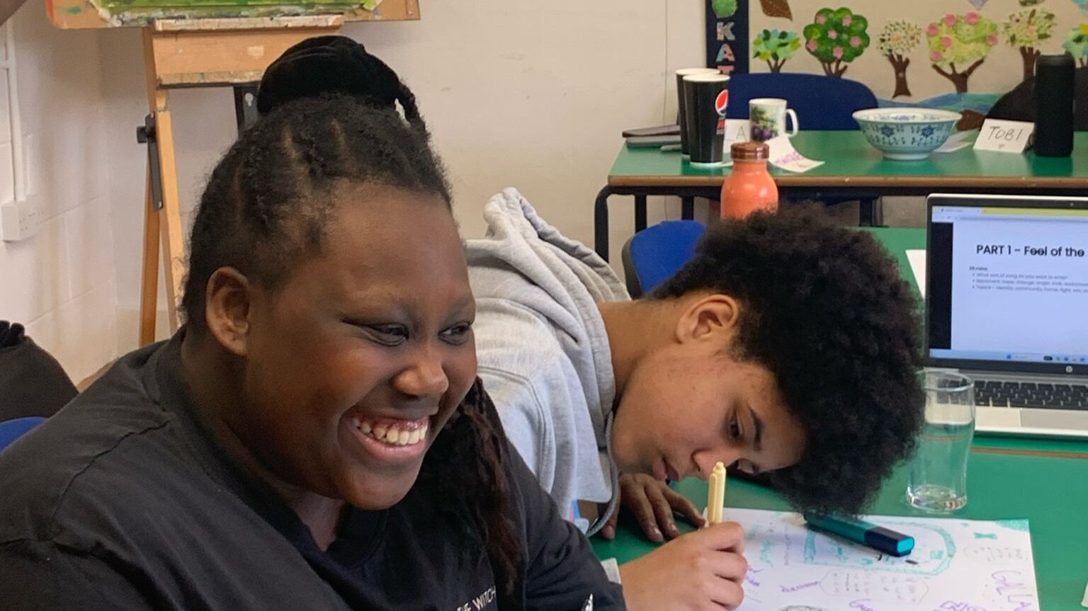 Two young black people drawing at a table. One leans over with a pen, the other is smiling broadly.