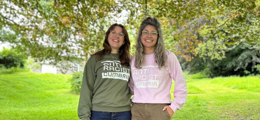 Two white women wearing glasses stand side by side under a tree. They are smiling broadly and both wear sweatshirts that read Anti Racist Cumbria.