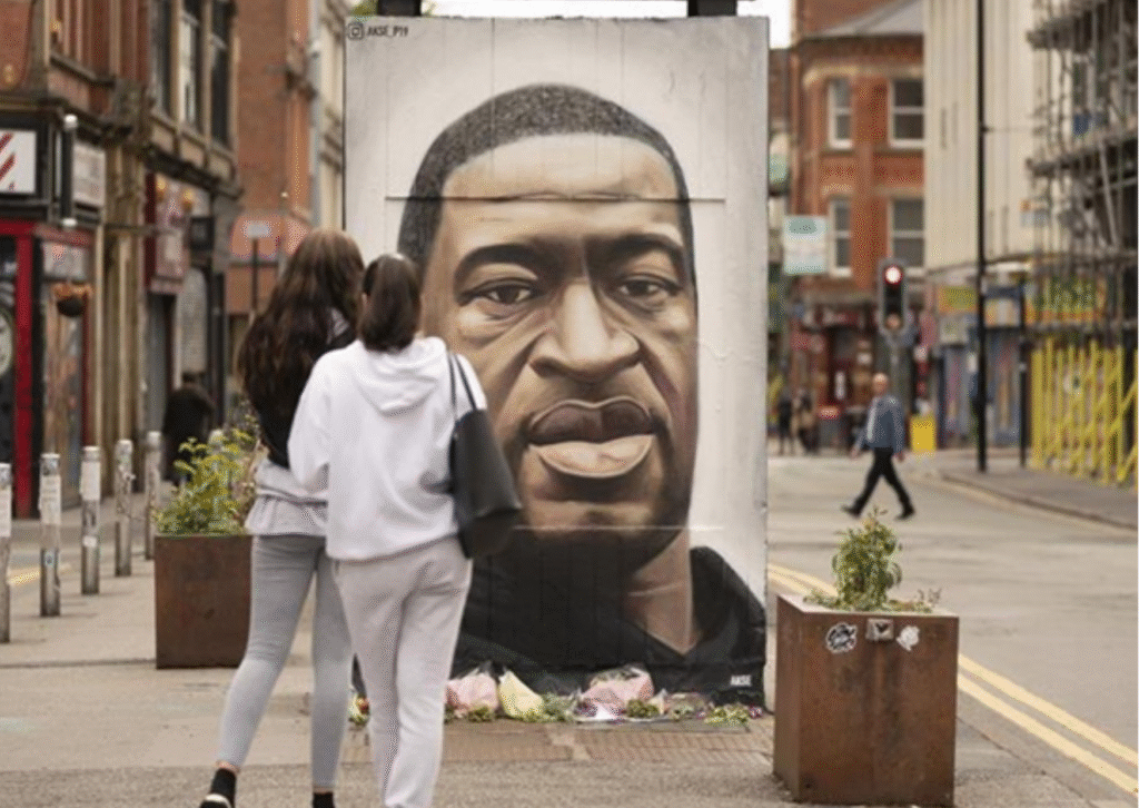 Image shows two Black women facing away from the camera as they look at a mural of a Black man (George Floyd). Flowers can be seen laid out in front of him.