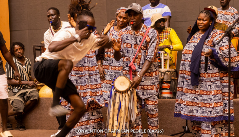 Image shows Black performers is beautiful, bright outfits singing and dancing. The picture exudes joy.